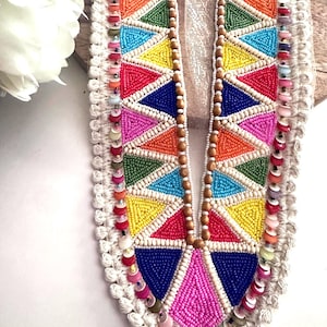 Op de afbeelding: Een kleurrijke kralenhalslijn met een geometrisch patroon van driehoeken. De halslijn is afgewerkt met witte franjes en heeft een houten achterkant.