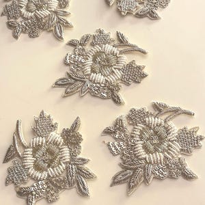 Op de afbeelding: Vijf zilveren bloemenapplicaties met ingewikkeld kralenwerk en pailletten. Elke applicatie heeft een centrale bloem omringd door gedetailleerde bladeren, perfect voor het verfraaien van kleding of knutselwerk.