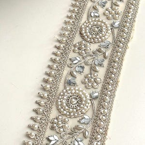 May include: A decorative trim featuring a floral design with faux pearls and silver-colored accents. The trim has a cream-colored base with a detailed pattern of pearls, silver leaves, and floral elements.