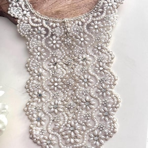 May include: A white beaded and sequined lace collar with a scalloped edge. The collar is made of pearls, sequins, and beads.