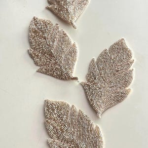 May include: Four silver beaded feather appliques on a white background. Each applique is shaped like a feather, with detailed beadwork creating the veins and edges. These decorative elements are suitable for craft projects, adding a touch of elegance.