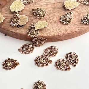 Può includere: Una collezione di decorazioni floreali in perline dorate, alcune con un retro marrone, sparse su una superficie bianca.