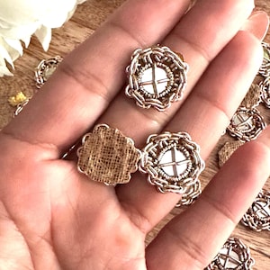 May include: Close-up of several small, round, silver-toned buttons with a cross design on a white background. The buttons have a chain-like border and are set on a burlap backing. The buttons are held in a hand.