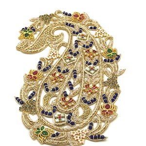 Könnte beinhalten: Eine paisley-förmige Applikation mit goldener Metallfadenstickerei, verziert mit kleinen blauen Perlen, klaren Kristallen und floralen Emaille-Motiven in Rot, Grün und Weiß. Das komplexe Design weist geschwungene Muster und feine Details auf.