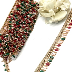 Può includere: Primo piano di finiture decorative con perline rosse, verdi e trasparenti. È visibile anche un bordo dorato con perline ovali rosse, verdi e rosa. Un fiore bianco è sullo sfondo. La finitura viene probabilmente utilizzata per abbellimenti.