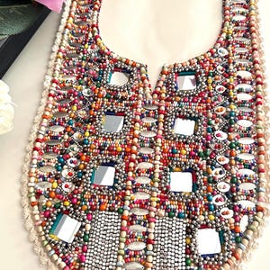 May include: A colorful beaded collar with square mirror accents. The collar features a variety of beads in shades of red, orange, yellow, green, and blue. The edges are trimmed with a cream-colored lace.