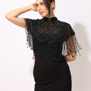 Model wearing a black embroidered beaded cape with scalloped edges and fringe. The sheer net base is hand-embellished with intricate embroidery and sparkling black beads, creating a statement bridal or evening accessory.