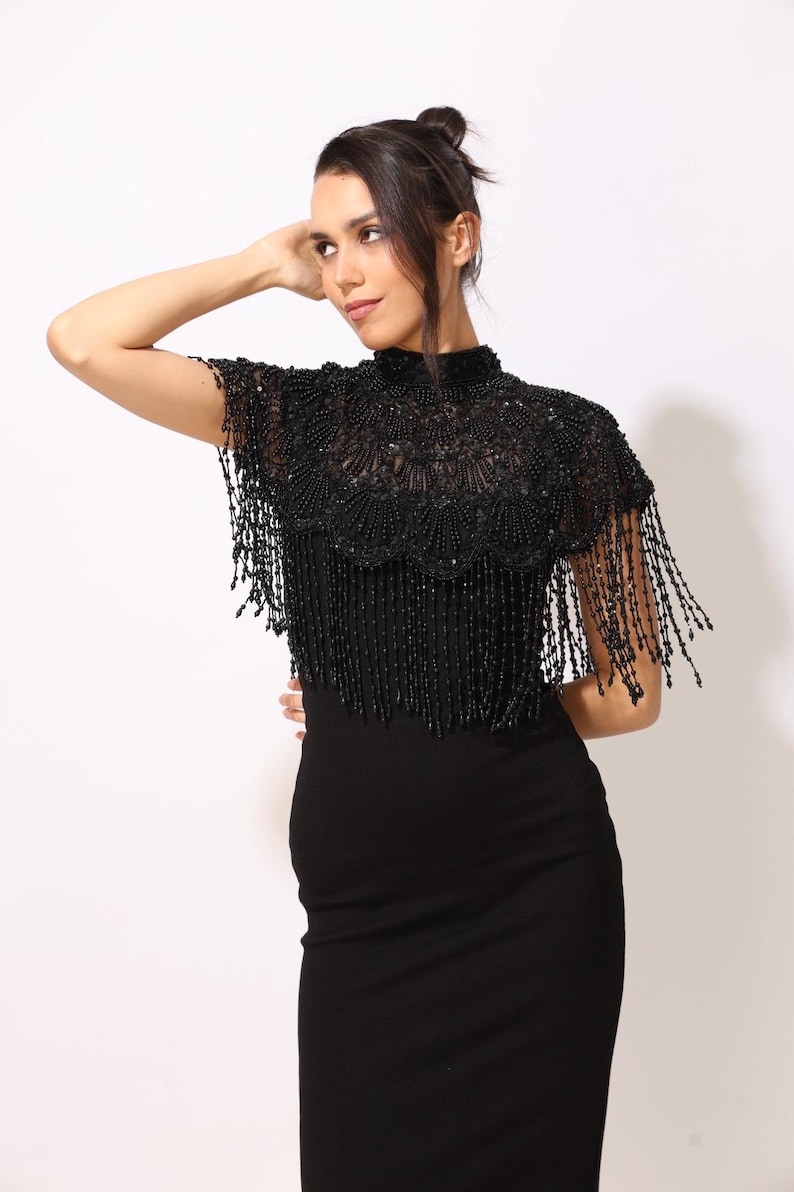 Model wearing a black embroidered beaded cape with scalloped edges and fringe. The sheer net base is hand-embellished with intricate embroidery and sparkling black beads, creating a statement bridal or evening accessory.