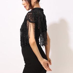 Model wearing a black embroidered beaded cape with scalloped edges and fringe. The sheer net base is hand-embellished with intricate embroidery and sparkling black beads, creating a statement bridal or evening accessory.