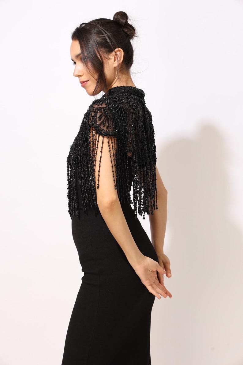 Model wearing a black embroidered beaded cape with scalloped edges and fringe. The sheer net base is hand-embellished with intricate embroidery and sparkling black beads, creating a statement bridal or evening accessory.