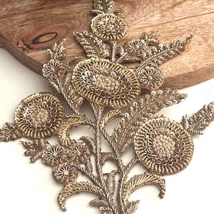 May include: Gold floral embroidered applique with intricate detailing. The applique features multiple flower shapes with beaded centers and delicate leaves. The design is made with gold thread and sequins, creating a shimmering effect. Perfect for embellishing garments or accessories.