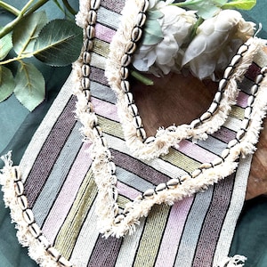 Puede incluir: Un ribete de cuentas a rayas con flecos y detalles de conchas. El ribete es en tonos de rosa, verde, blanco y marrón.