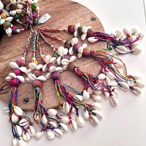 May include: A collection of colorful tassels with white seashells. Each tassel is made with a variety of colorful threads and has a cluster of white seashells at the end. The tassels are arranged on a wooden surface.
