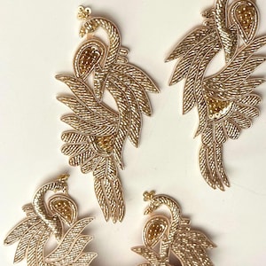 May include: Four gold embroidered peacock appliques. Each applique features a detailed design with beaded accents and a textured, feathered appearance. The appliques are arranged on a white surface, showcasing their intricate craftsmanship.