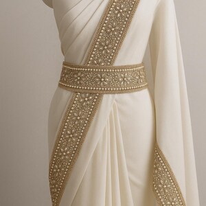 May include: Ivory-colored saree with intricate gold embroidery and pearl accents. The saree features a decorative belt with floral patterns and a matching border along the edges. The fabric drapes elegantly, showcasing the detailed craftsmanship of the garment.