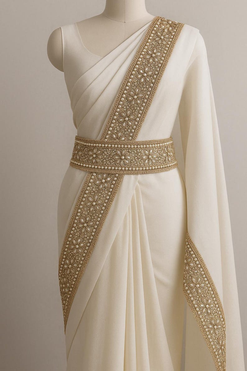May include: Ivory-colored saree with intricate gold embroidery and pearl accents. The saree features a decorative belt with floral patterns and a matching border along the edges. The fabric drapes elegantly, showcasing the detailed craftsmanship of the garment.