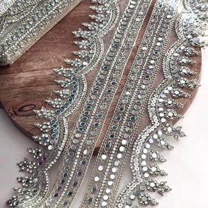 May include: Silver beaded trim with reflective mirror accents. The trim features a delicate, ornate design with small, round mirrors and sparkling beads. The trim is ideal for embellishing garments, accessories, or home decor projects.