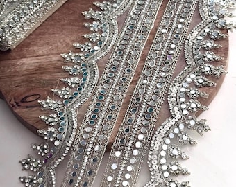 May include: Silver beaded trim with reflective mirror accents. The trim features a delicate, ornate design with small, round mirrors and sparkling beads. The trim is ideal for embellishing garments, accessories, or home decor projects.