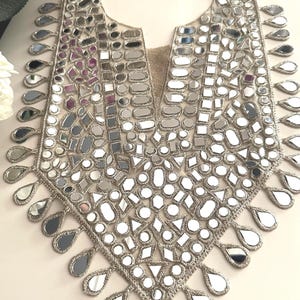 May include: A silver-coloured, decorative collar with a V-neck design. The collar is embellished with numerous small, irregularly shaped mirrors and teardrop-shaped mirror pendants along the edges. The base material appears to be a woven fabric.