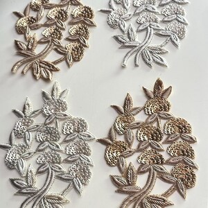 May include: Four embroidered floral appliques in gold and silver. Each applique features a cluster of small, round embellishments and detailed leaf accents. The appliques are arranged on a white surface, showcasing their intricate design.