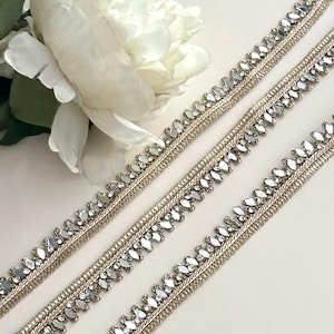 May include: Close-up of three decorative silver trim ribbons with clear, reflective embellishments. The ribbons are arranged diagonally, with a white flower in the upper left corner. The trim has a woven design.