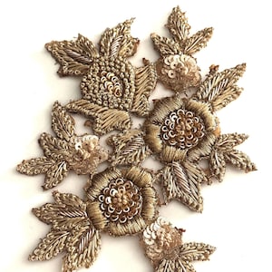 May include: An ornate, gold-colored floral applique with intricate embroidery and sequin details. The design features multiple flower and leaf shapes, creating a textured, three-dimensional effect. Ideal for embellishing garments or craft projects.
