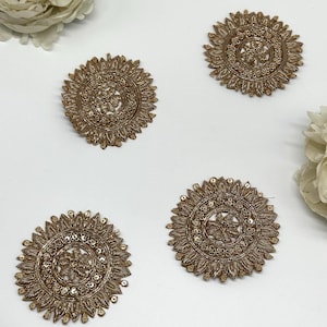 Può includere: Quattro applicazioni ricamate circolari color oro con intricati motivi floreali e a foglie. Ogni applicazione presenta un disegno centrale circondato da un bordo dettagliato e piccole paillettes. Le applicazioni sono disposte su una superficie bianca.