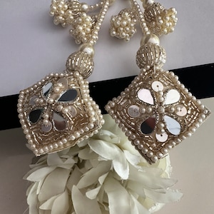 Puede incluir: Dos borlas blancas de cuentas con detalles de flores de espejo. Las borlas están hechas de cuentas blancas y tienen una forma cuadrada con un diseño de flor de espejo en el centro. Las borlas cuelgan de un fondo blanco.