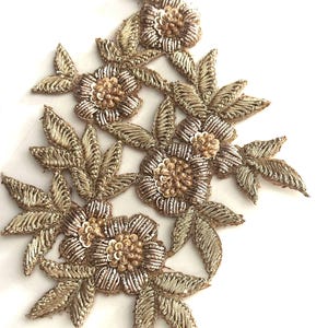 Puede incluir: Aplicación floral bordada en tonos dorados y plateados. El diseño presenta múltiples flores con detalles de pétalos y centros con cuentas, rodeadas de hojas cosidas intrincadamente. Ideal para embellecer prendas o proyectos de manualidades.