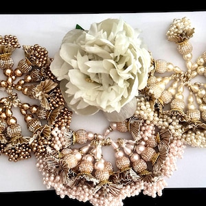 Può includere: Una collezione di nappine di perline dorate e rosa cipria con un fiore di seta bianco al centro. Le nappine sono fatte di piccole perline e hanno un design a frangia.