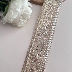 Roll of intricate gold pearl and mirror lace ribbon, displayed with white flowers for wedding, costume, or ethnic garment use.