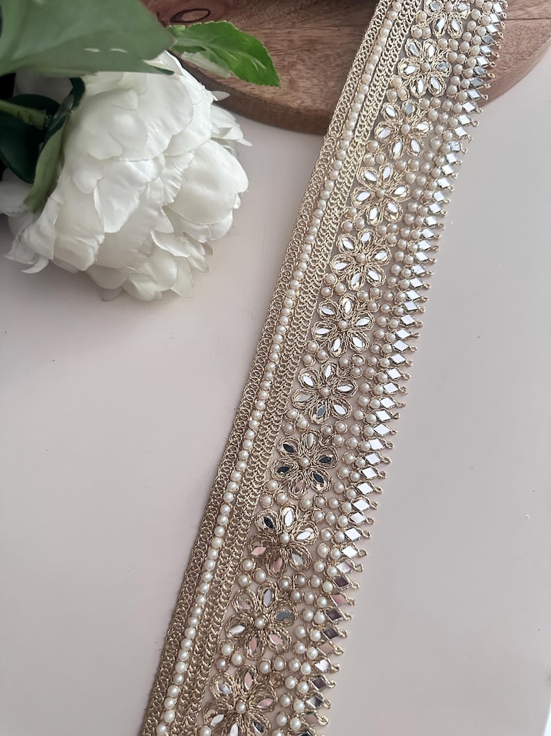 Roll of intricate gold pearl and mirror lace ribbon, displayed with white flowers for wedding, costume, or ethnic garment use.