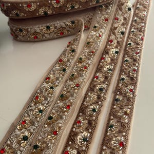 以下が含まれることがあります： ベージュ色のファブリックトリムで、複雑な金色の刺繍、小さな白いビーズ、赤と濃い緑の装飾用スパンコールが施されています。トリムは巻かれたロール状と数本の直線状に配置されており、全体に繰り返される花柄のデザインが見られます。