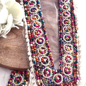 May include: A colourful beaded trim with a V-shaped neckline, featuring a variety of beads in different shapes and sizes, including wooden beads, glass beads, and pearl-like beads. The trim is accented with a fringe of white yarn.