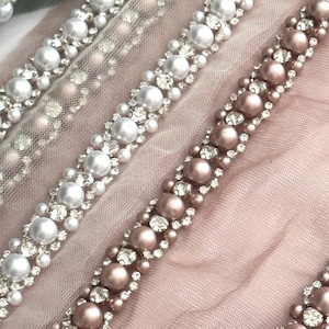 May include: Two rows of beaded trim featuring pearl-like beads and rhinestones. The trim is displayed on a light pink fabric.