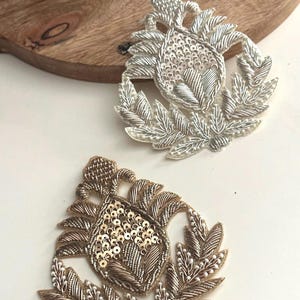 May include: Two embroidered patches with a floral design. One is silver with silver sequins, and the other is gold with gold sequins. Both have a leaf-like border and are on a white surface.