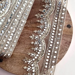 May include: Close-up of silver decorative trim with reflective mirror accents. The trim is arranged on a wooden surface. The trim features intricate patterns and is likely used for embellishing garments or crafts.