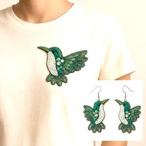 Op de afbeelding: Een paar groene kolibrie-oorbellen en een bijpassende broche. De oorbellen en de broche zijn gemaakt van groene en witte kralen en pailletten.