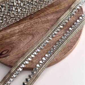 Puede incluir: Primer plano de un adorno decorativo con acabado dorado metálico. El adorno presenta un patrón repetitivo de acentos espejados en forma de lágrima y pequeños brillantes transparentes. El adorno se muestra sobre una superficie de madera.