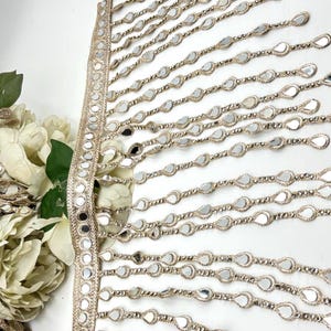 May include: Gold-coloured decorative trim featuring a woven base and dangling teardrop-shaped mirrored accents. The trim has small, round mirrors along the edge. Suitable for embellishing clothing or craft projects.