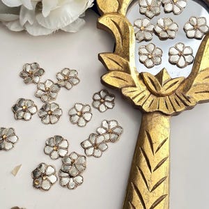 May include: A collection of silver and gold embroidered flower embellishments. The flowers are arranged on a white surface and in a gold mirror.
