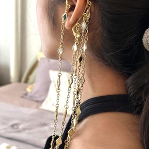 May include: Gold-coloured ear cuff with dangling chains. The ear cuff has diamond-shaped charms in black, white, and gold. The chains cascade down, creating a layered effect. The jewellery is worn on the ear.