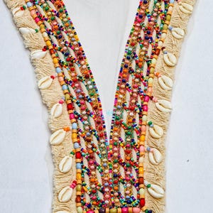 Peut inclure: Un col en V orné de perles colorées, de coquillages et de franges. Le design présente une variété de petites perles rondes dans les tons de rouge, orange, jaune, vert, bleu et rose. Des coquillages blancs sont parsemés le long des bords.