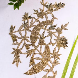 Puede incluir: Diseño de pavo real bordado en oro con detalles florales. El intrincado bordado presenta hilo metálico y lentejuelas, creando un patrón detallado y ornamentado. El diseño se encuentra sobre un fondo blanco con hojas verdes.