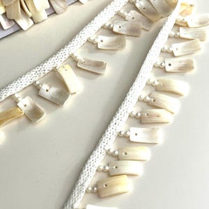 Puede incluir: Borde blanco con un borde de cuentas y una franja de piezas rectangulares blancas con forma de concha.