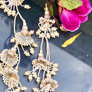 May include: A pair of white pearl and silver tassel earrings with a floral design. The earrings are hanging from a branch with a pink flower.