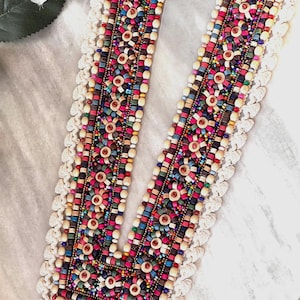 Op de afbeelding: Een kleurrijke kralenbies met een V-hals, met een mix van houten en glazen kralen in verschillende kleuren en maten. De bies is afgewerkt met een wit gecrochete franje.