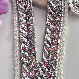 May include: A white crochet trim with a silver and multi-colored beaded design. The beaded design is in a chevron pattern and is attached to a sheer fabric.