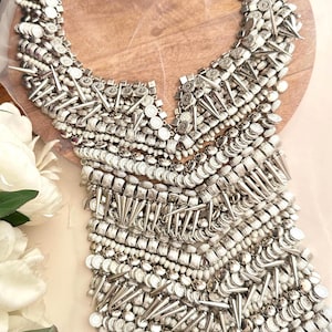 以下が含まれることがあります： 様々な大きさの金属ビーズとスパイクで構成された、大きく複雑なデザインの銀色のネックレス。ネックレスは木製の表面に置かれています。