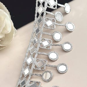 Puede incluir: Borde plateado con un patrón repetido de espejos en forma de diamante y espejos redondos. El borde tiene una franja de espejos redondos.